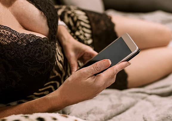Reife Frau in Dessous mit Handy auf dem Bett